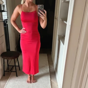 Röd långklänning med smala axelband - Snygg röd långklänning med smala axelband och öppen rygg. Klänningen har en tight passform och slits baktill. Perfekt för dig som vill sticka ut med en enkel men stilren look.
