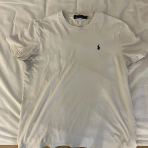 Vit t-shirt från Polo Ralph Lauren - Vit t-shirt från Polo Ralph Lauren med klassisk rund hals och korta ärmar. T-shirten har den broderade loggan på bröstet och är gjord i mjuk bomull. Passformen är normal och stilen är clean och enkel. Perfekt för dig som gillar stilren och fräsch stil. Hör ev dig vid minsta fundering😊