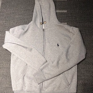 Grå hoodie från Ralph Lauren - Snygg grå hoodie från Ralph Lauren med dragkedja och klassisk logga broderad på bröstet. Tröjan har huva, långs ärmar och två fickor framtill. Perfekt för en chill och stilren look. Finns små slitningar där nere på hoodien och finns ett litet hål i armhålan, men det är ställen man inte tittar på.