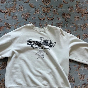 Vit SpongeBob sweatshirt från Divided - Vit sweatshirt med svart SpongeBob-tryck och texten 'Dare to be square' på bröstet. Tröjan har rund hals, långa ärmar och är gjord i mjuk bomull och polyester. Perfekt för dig som gillar coola prints och vill sticka ut lite extra.