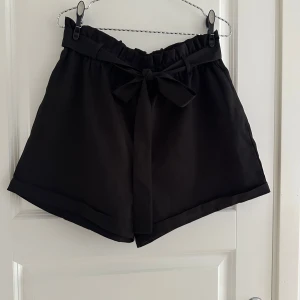 Svarta shorts med knytband - Stilrena svarta shorts med hög midja och brett knytband i midjan. De har en lös passform och är tillverkade i ett mjukt, lätt tyg som ger en skön känsla. Perfekta för varma dagar när du vill ha något både bekvämt och snyggt.
