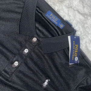 Svart pikétröja från Ralph Lauren - Klassisk svart pikétröja från Ralph Lauren med vit broderad logga på bröstet. Tröjan har krage, tre knappar framtill och korta ärmar. Perfekt för en clean och stilren look. Materialet är mjukt och bekvämt, troligtvis bomull.