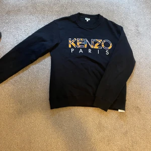 Svart Kenzo sweatshirt med broderi - Svart sweatshirt från Kenzo med stort broderat logotyptryck i orange, lila och blått på bröstet. Tröjan har rund halsringning och långa ärmar. Perfekt för dig som vill sticka ut med en snygg streetstyle-look.
