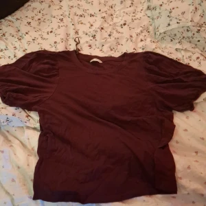 Vinröd puffärm blus i ekologisk bomull - Snygg vinröd blus från Cubus i ekologisk bomull, med puffiga korta ärmar och rund halsringning. Perfekt för dig som gillar en chill men ändå trendig look. Materialet är mjukt och skönt mot huden och färgen är djup och mörk. Passar dig som vill sticka ut lite extra.