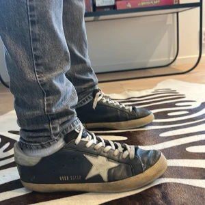 Svarta GGDB Superstar sneakers - Svarta GGDB Superstar sneakers med vit stjärndetalj på sidan och beige slitna kanter. Skorna har snörning och platt sula i gummi, samt rund tå. Ovandelen är i skinn och hälen är vit. Perfekt för dig som gillar en cool och avslappnad streetstil.