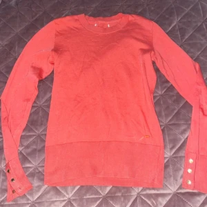Korallfärgad stickad tröja holly&whyte, S - Snygg korallfärgad stickad tröja från holly&whyte med rund hals och långa ärmar. Tröjan har coola guldfärgade knappar vid ärmsluten och en liten metallbricka med logga vid nederkanten. Perfekt för dig som gillar stilrena detaljer och mjuka material.