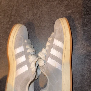 Adidas Campus grå mocka sneakers - Snygga Adidas Campus sneakers i ljusgrå mocka med klassiska vita ränder och vit sula. Skorna har rund tå, platt sula och breda vita snören. Perfekta för dig som gillar retrostil och vill ha något som funkar till det mesta.