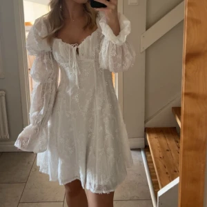 Studentklänning NA-KD - Superfin vit kort klänning med puffärmar och broderade blommor. Klänningen har snörning vid bysten och är tillverkad i ett tunt, luftigt material. Den har dubbla lager med tyg ner till, så den är inte genomskinlig!! Perfekt för student🌼endast använd vid ett tillfälle 