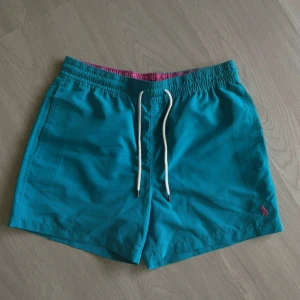 Turkosa badshorts Polo Ralph Lauren - Slim fit turkosa badshorts från Polo Ralph Lauren med vit snörning i midjan och rosa insida. Klassisk design med broderad logga i rosa på benet. Tillverkade i 100% polyester och har meshfoder för extra komfort.