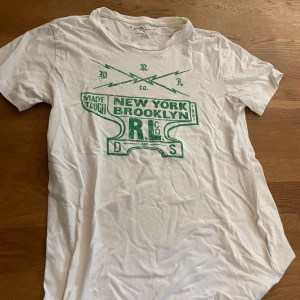 Vit t-shirt Ralph Lauren Brooklyn - Snygg vit t-shirt från Denim & Supply Ralph Lauren med grön Brooklyn/New York-print framtill. Klassisk rund hals och korta ärmar. Tillverkad i mjuk 100% bomull, perfekt för chill dagar. Passformen är normal och trycket ger en cool streetkänsla.