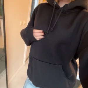 Svart oversized hoodie med ficka - En svart hoodie med stor känguruficka och snörning i huvan. Tröjan är oversized och har långa ärmar, perfekt för chill dagar. Mjuk insida och klassisk look som funkar till jeans eller mjukisbyxor.