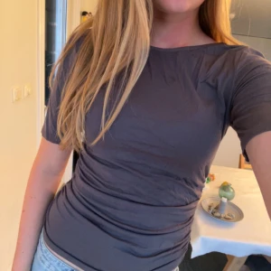 Mörkgrå t-shirt med öppen rygg - Snygg mörkgrå t-shirt med korta ärmar och rund hals. T-shirten har cool snörning längs sidan som ger en unik look och sitter tight mot kroppen. Baktill är det öppen rygg men inte hela vägen (fråga om bild om du undrar.). Materialet känns som mjuk bomull och passar perfekt till jeans eller kjol. Enkelt att styla för en chill vibe.