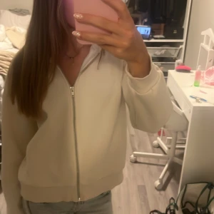 Beige hoodie med dragkedja - Säljer en stilren beige hoodie med dragkedja framtill och huva. Tröjan har långa ärmar, ribbade muddar och en mjuk insida. Perfekt för dig som gillar en enkel och clean look. Passar till jeans eller mjukisbyxor.