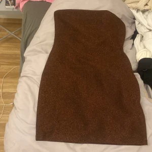 Brun glittrig tubklänning H&M Divided - Säljer en brun glittrig tubklänning från H&M Divided i storlek S. Klänningen är kort och figurnära med snygg glittereffekt, perfekt för att sticka ut. Axelbandslös modell som ger en clean och trendig look.