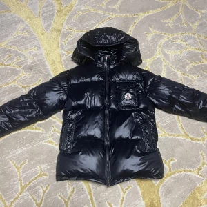 Svart Moncler pufferjacka med huva - Snygg svart pufferjacka från Moncler med glansig finish, huva och dragkedja framtill. Jackan har en bröstficka med lock och Moncler-logga, samt två sidofickor. Perfekt för kalla dagar och riktigt street. Fodrad och dunfylld för extra värme.