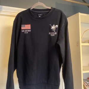 Polo club tröja - Svart sweatshirt från Vinson Polo Club med broderad logga och amerikansk flagga på bröstet. Klassisk rund hals och långa ärmar. Mjuk bomullskänsla och relaxed fit, perfekt till jeans eller joggers. Säljer för att den är för liten för mig