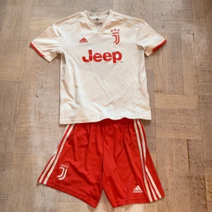 Juventus fotbollströja - Juventus matchtröja från Adidas i vit med röda detaljer och tryck. Kortärmad med V-ringad krage, tillverkad i lätt och ventilerande Climalite-material. Rött Juventus-emblem och Jeep-logga på bröstet. Säljes med röda shorts med vita ränder och Juventus-logga.