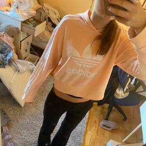 Croppad rosa hoodie från Adidas - Säljer en croppad hoodie från Adidas i ljusrosa med vit logga och klassiska ränder på ärmarna. Tröjan har huva, långa ärmar och är i storlek 164 men passar xxs-s