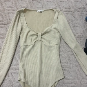 Beige ribbad långärmad topp - Snygg beige ribbad topp med lång ärm och djup urringning. Toppen har en knutdetalj framtill som ger en cool twist. Perfekt att styla med jeans eller kjol för en trendig look.