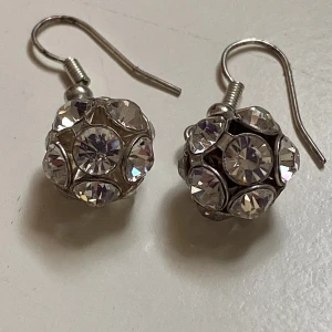 Glittriga silverörhängen med kristaller - Säljer ett par snygga örhängen i silverfärg med rund form och massor av glittrande, klara kristaller. De har krokar för enkel användning och bollarna är täckta av stenar som fångar ljuset. Perfekta för dig som vill sticka ut med lite extra bling.