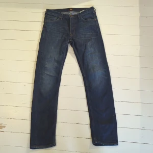 Lee Daren Button Fly jeans blå W31 L34 - Snygga Lee Daren Button Fly jeans i mörkblå tvätt med klassisk femficksdesign och bruna Lee-detaljer. Jeansen har rak passform och är gjorda av ekologisk bomull. Perfekt för dig som gillar stilrena och tidlösa jeans med knappgylf.
