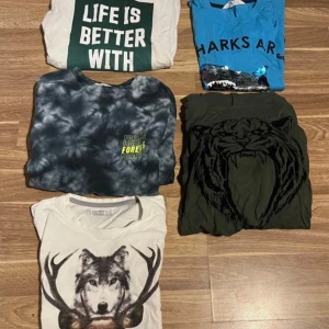 Fem coola t-shirts med tryck - Fem t-shirts med olika tryck: vit med texten 'Life is better with', blå med hajmotiv, svart/grå batik med neontext, mörkgrön med stor tigertryck och vit med varg och horn. Alla har rund hals och korta ärmar, perfekta för dig som gillar unika prints och djurmotiv.