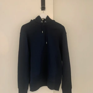 Mörkblå hoodie från J.Lindeberg - Snygg mörkblå hoodie från J.Lindeberg med klassisk känguruficka och justerbar huva med snörning. Tröjan är i mjuk bomull och har långa ärmar, perfekt för chill dagar eller när du vill ha en clean look.