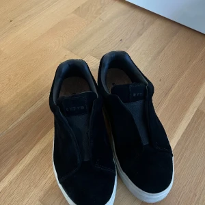 Svarta Eytys sneakers i mocka - Säljer ett par svarta Eytys sneakers i mocka med vit platt sula och lila detalj baktill. Skorna har en minimalistisk design utan snörning och en rund tå. 