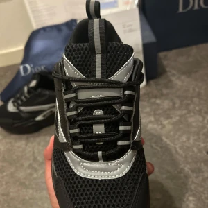 Svarta D1or sneakers med silverdetaljer - Snygga svarta sneakers från D1or med coola silverdetaljer och meshpaneler. Skorna har rund tå, platt sula och snörning framtill. Märket syns tydligt på sidan och innersulan. Perfekt för dig som gillar exklusiv streetstyle och vill sticka ut med lyxiga detaljer.