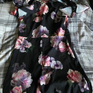 Blommig svart klänning från Lipsy - Supersnygg svart klänning från Lipsy London med stora blommor i rosa, lila och vitt. Klänningen har kort längd, v-ringad rygg och axelbandslös design. Perfekt för dig som gillar färgstarka och romantiska plagg med coola detaljer.