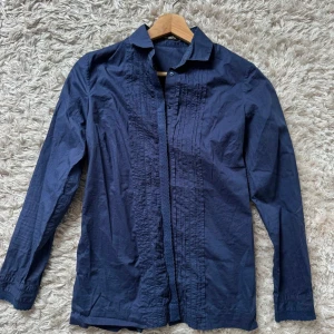 Mörkblå skjorta med veckade detaljer - Snygg mörkblå skjorta med lång ärm och diskreta veckade detaljer framtill. Klassisk krage och knäppning framtill, perfekt för en stilren look. Skjortan är i ett lätt och bekvämt material som känns skönt mot huden.