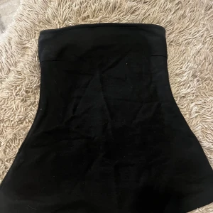 Svart tubetop från Gina Tricot - Enkel och stilren svart tubetopp från Gina Tricot. Toppen har en tight passform upptill och lätt utsvängd nertill, vilket ger en snygg siluett. Perfekt att matcha med jeans eller kostymbyxor