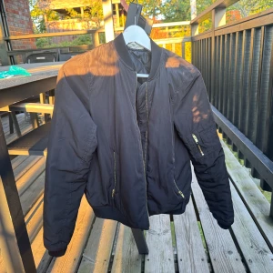 Svart bomberjacka med guldiga detaljer - Säljer en svart bomberjacka med guldiga dragkedjor och klassisk ribbad krage. Jackan har fickor med dragkedja och en extra ficka på ärmen