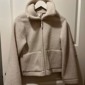 Beige teddyjacka från H&M  - Mysig beige teddyjacka från H&M i storlek XS. Jackan har hög krage, dragkedja framtill och två stora fickor. Perfekt för kalla dagar och ger en chill vibe med sin fluffiga look.