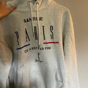 Ljusgrå hoodie med Paris-tryck - Säljer en ljusgrå hoodie från Santigie med stor Paris-tryck på bröstet och texten 'Ce n'est pas fini'. Hoodien har snörning i huvan, känguruficka och detaljer i blått och rött. 