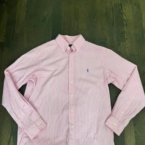 Rosa randig skjorta från Polo Ralph Lauren - Snygg rosa och vit randig skjorta från Polo Ralph Lauren med klassisk button-down krage och broderad blå logga på bröstet. Skjortan har långa ärmar och vita knappar framtill. Perfekt för dig som gillar stilrena och fräscha plagg. Öppen för byten och går gärna ner i pris vid en snabb affär så våga släng ett bud