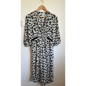 Blommig långklänning från Gerard Darel - Supersnygg långklänning från Gerard Darel i vitt med svart blommönster och två fickor. Djup v-ringning med hyskknäppning, trekvartsärmar och markerad midja med resår. 