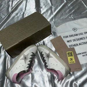 Golden Goose Superstar sneakers vit/rosa - Golden Goose Superstar sneakers i vitt skinn med grå stjärndetalj på sidan, grå snörning och ljusrosa hälparti. Platt sula och rund tå. Märkets logga syns på hälen och tungan. Snygg kontrast mellan färgerna och cool vintagekänsla.