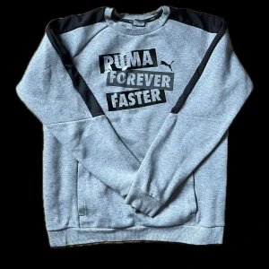 Grå sweatshirt från Puma - Säljer en grå sweatshirt från Puma med texten 'Puma Forever Faster' i svart på framsidan. Tröjan har svarta detaljer på axlarna och ärmarna samt ribbade muddar vid ärmslut och nederkant. Perfekt för en sportig look! OBS Liten skada vid höger arm