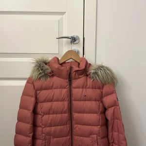 dunjacka från Tommy Hilfiger - Säljer en snygg rosa dunjacka från Tommy Hilfiger med pälskantad huva. Jackan har en dragkedja framtill och är perfekt för kyliga dagar. Den har en klassisk design med märkets logga på bröstet och ena armen. Priset kan diskuteras. 💗