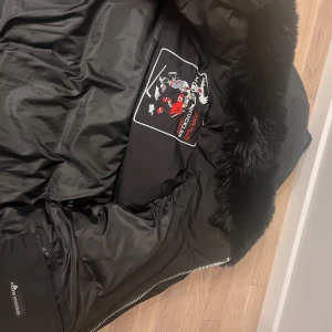 Svart pufferjacka med pälsdetaljer - Säljer en snygg svart pufferjacka från Moose Knuckles. Den har en fluffig pälsdetalj på huvan och coola dragkedjor. Perfekt för kalla vinterdagar! Jackan är i mycket bra skick och har en broderad logga på insidan. Passar både till vardags och fest. Går inte att stänga med zip bara med knapparna 