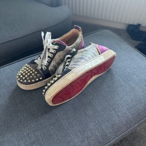 Glittriga sneakers med nitar - Unika sneakers med glittrigt mönster och guldnitar på tån. Skorna har en mix av färger som silver, svart och lila, med vita snören och en röd sula. De är bara lite smutsiga men inget sönder.