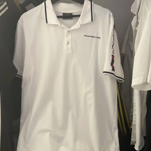 Vit pikétröja från Porsche - Säljer en stilren vit pikétröja från Porsche med svarta detaljer och text på ärmen. Tröjan har en klassisk krage med knappar och märkeslogga på bröstet. Perfekt för en sportig look! Säljer den för 900 nypris 1100kr