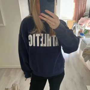 Mörkblå sweatshirt med text - Säljer en mörkblå sweatshirt med texten 'ATHLETIC' i vitt på framsidan. Tröjan har en avslappnad passform och långa ärmar, perfekt för en casual look. Passar bra till jeans eller leggings.