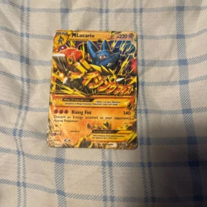 Pokémonkort Mega Lucario EX - Säljer ett Pokémonkort med Mega Lucario EX. Kortet har en kraftfull design med Lucario i en dynamisk pose och japansk text. Det är ett samlarkort med 220 HP och attacken Rising Fist. Perfekt för samlare eller Pokémon-entusiaster!