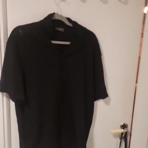 Svart kortärmad skjorta från H&M - Säljer en svart kortärmad skjorta från H&M med regular fit. Skjortan har ett diskret mönster och knappar framtill. Perfekt för en stilren look.