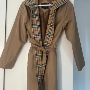 Beige trenchcoat från Burberry - Snygg beige trenchcoat från Burberry med klassiskt rutigt foder. Jackan har ett bälte i midjan och långa ärmar. Perfekt för en stilren look, dock lite fläckar. Men har inte försökt tvätta bort dem.