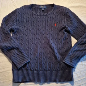 Mörkblå stickad tröja från Ralph Lauren - Säljer en stilren mörkblå stickad tröja från Ralph Lauren med kabelstickat mönster. Tröjan har en broderad röd logotyp på bröstet och långa ärmar med ribbade muddar. Perfekt för en klassisk look.