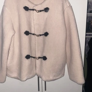 Beige teddyjacka från Gina Tricot - Mysig beige teddyjacka från Gina Tricot med svarta detaljer. Jackan har långa ärmar och stängs med tre svarta spännen framtill. Perfekt för kyliga dagar.