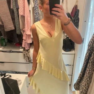 Gul ärmlös klänning med volang - Snygg gul klänning med v-ringning och volangdetaljer. Den är ärmlös och har en öppen rygg, perfekt för sommarens festligheter. Klänningen har en elegant och luftig design. Klänningen är helt ny med lappen kvar! Den kostar ju runt 600kr i nypris